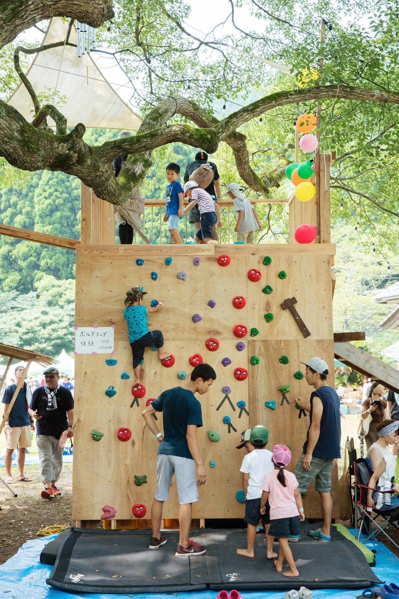 Children and adults climb a wooden wall with colorful holds beneath a large tree at an outdoor festival.