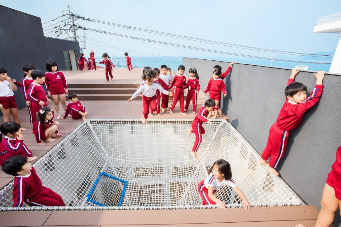 Children in red uniforms playing on a rooftop recreation area with netting and bay views.