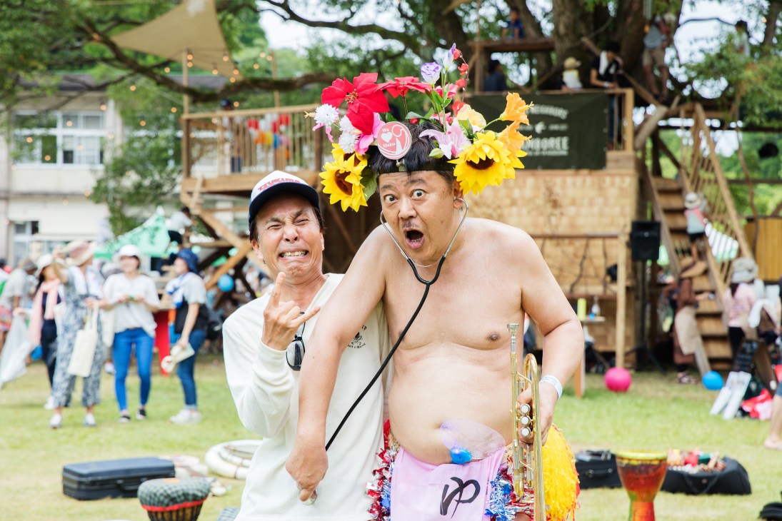 A shirtless musician wearing a floral headdress plays saxophone at an outdoor festival with crowd and treehouse visible in background.
