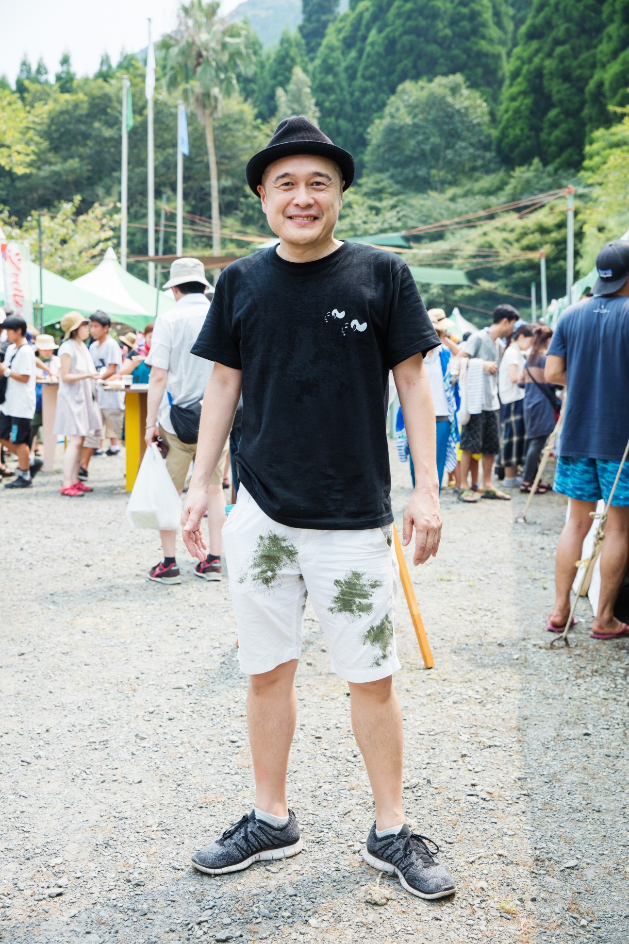 DJ Hiroshi Kawanabe smiling at the Good Neighbors Jamboree with festival crowds and forested mountains in the background.