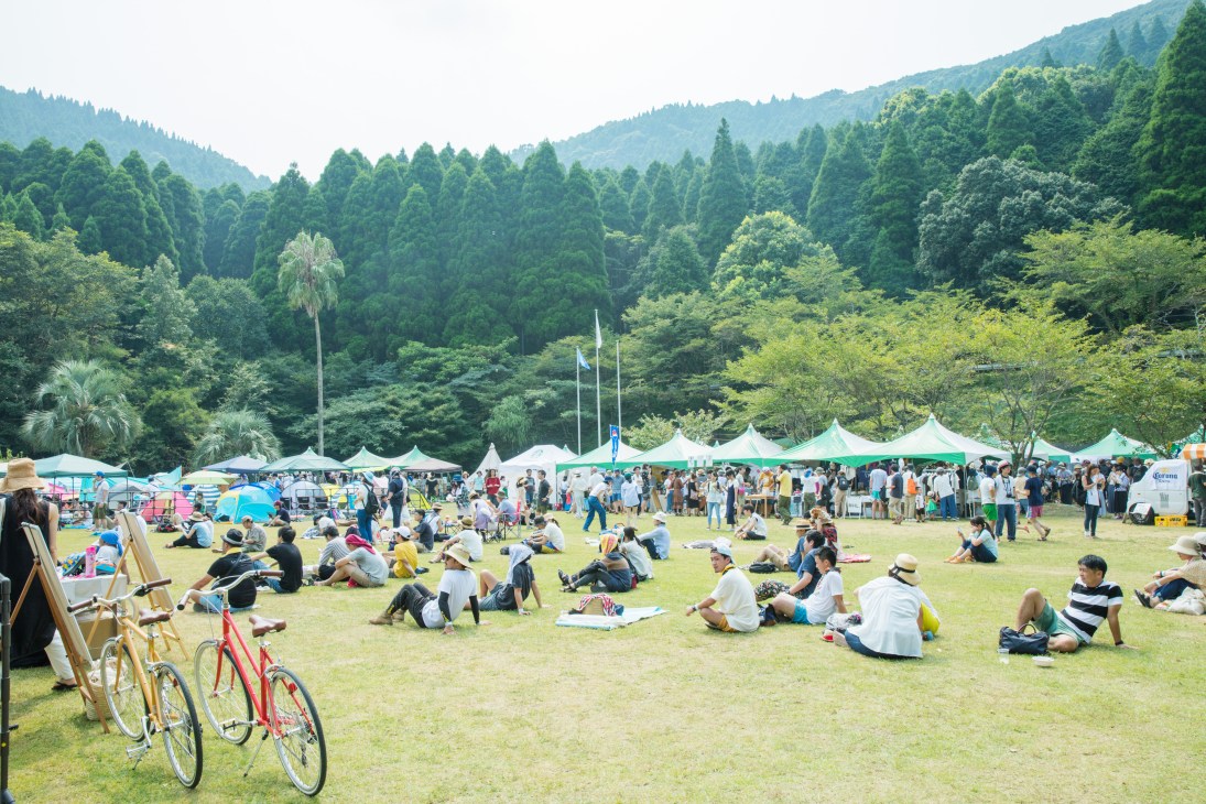 Aerial view of the Good Neighbors Jamboree festival grounds with white tents, visitors on grass, and forested mountains in background.