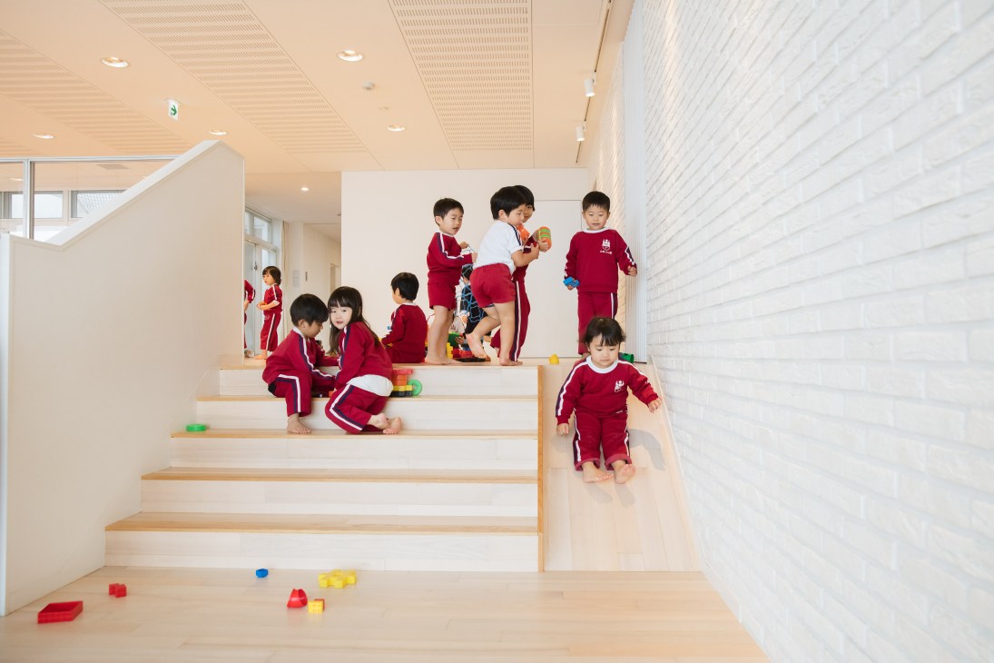 Young children in red uniforms playing on and around white stairs with wooden treads in a bright, minimalist interior space.