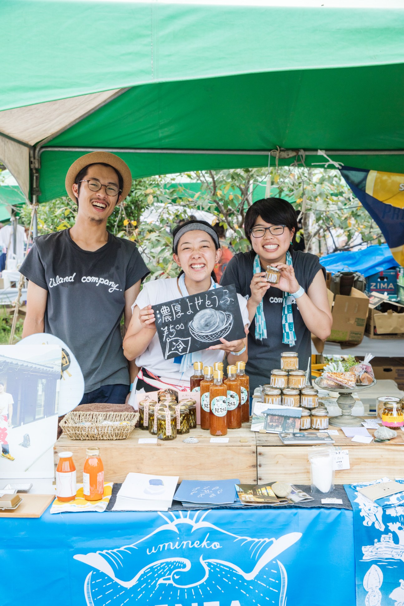 Three smiling vendors at Island Company stand under a green tent displaying jars of food products and local goods.