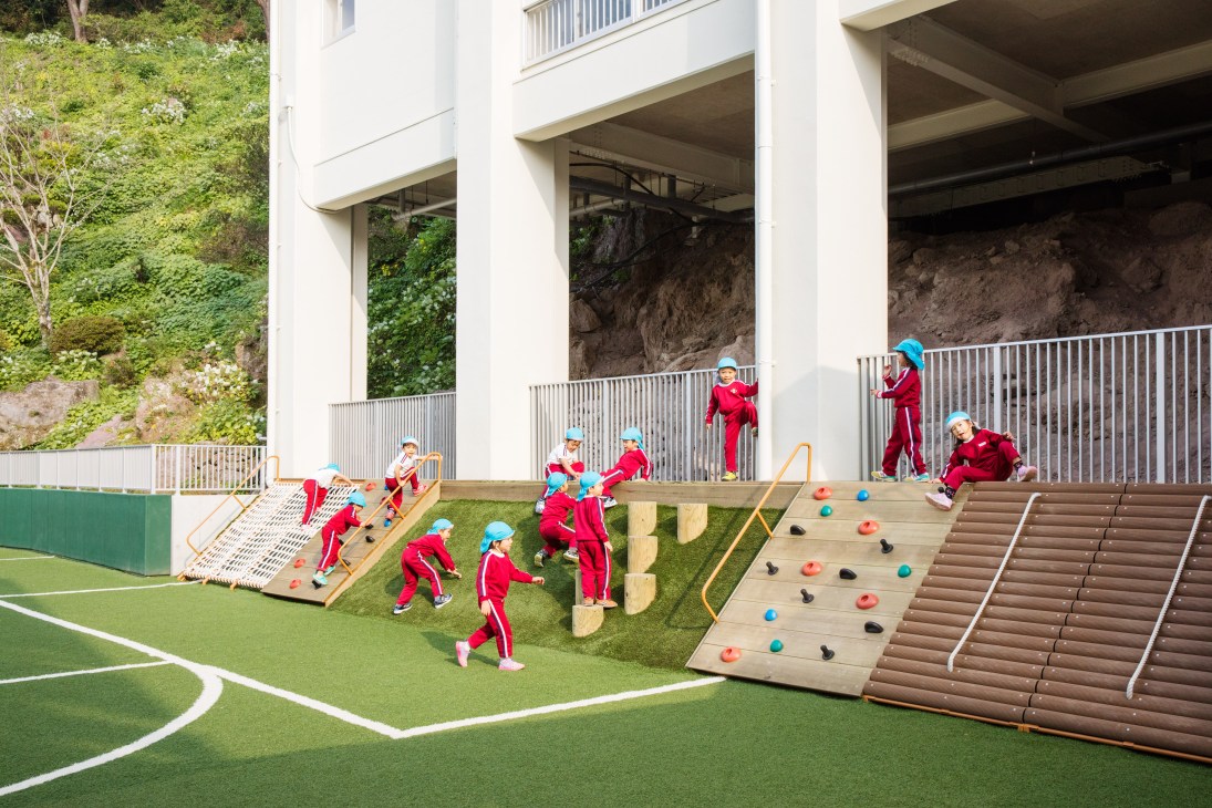 Children in red outfits play on climbing structures and ramps at Obama Kodomoen kindergarten in Japan.