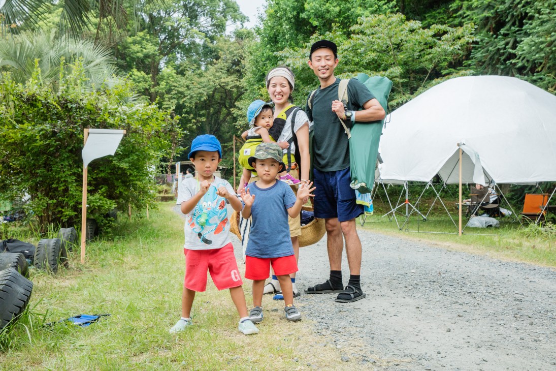 A young family with two children and two adults stand on a path surrounded by green forest and camping tents at an outdoor event.