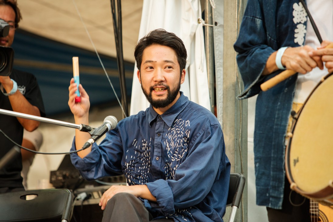 A bearded man in a blue patterned shirt sits at a microphone, smiling while performing with musicians holding instruments beside him.