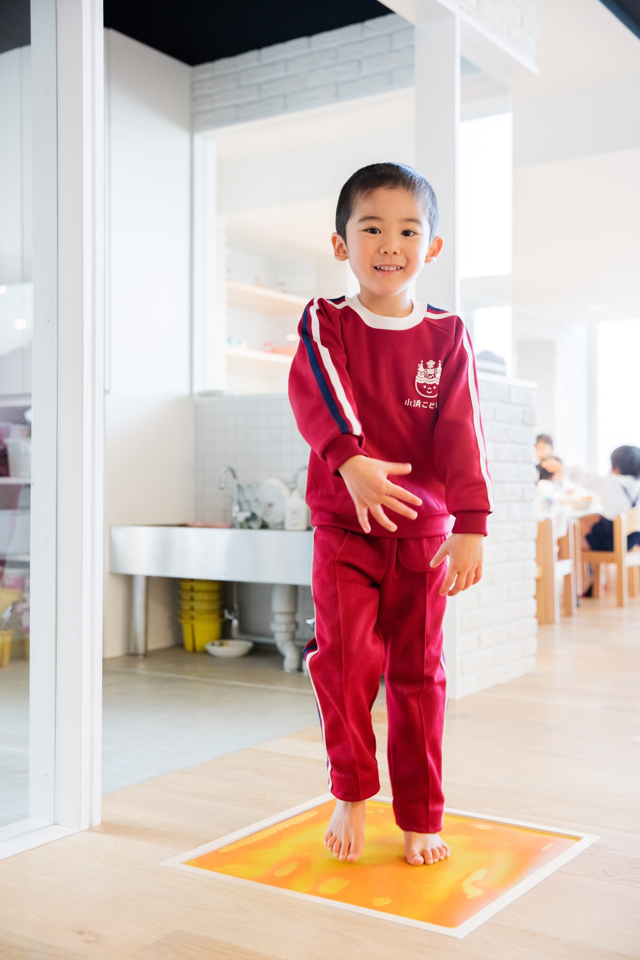 Young child in red school uniform standing on a color-changing floor section in a bright, modern nursery.