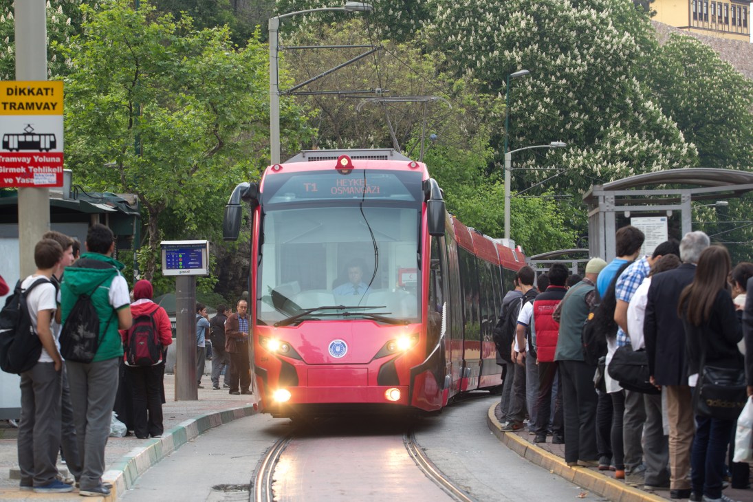 A red Silkworm tram carries passengers on Bursa's street surrounded by trees and waiting commuters.
