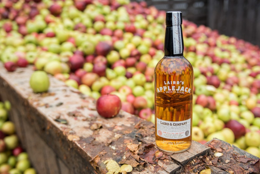 Bottle of Laird's Applejack brandy on wooden surface with apples in background orchard.