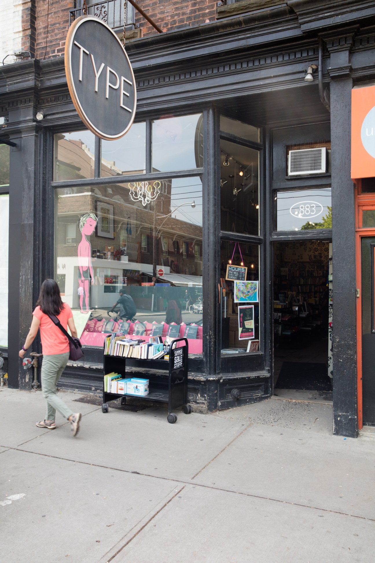 Type bookstore storefront with circular sign and woman walking past with books displayed in window
