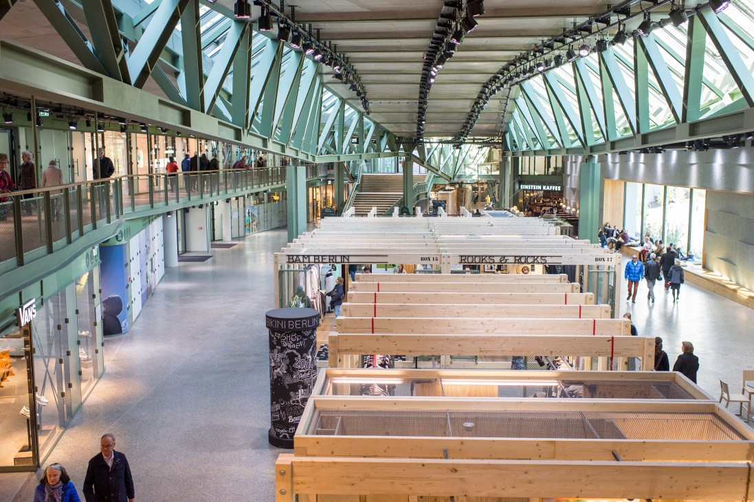 Interior gallery level of Bikini Berlin showing wooden pop-up booths with glass storefronts, green steel beams, and skylights.