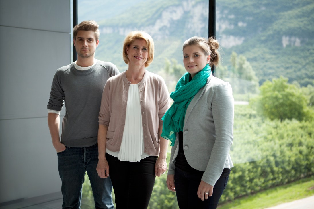 Three people posing indoors with a mountainous valley landscape visible through windows behind them.
