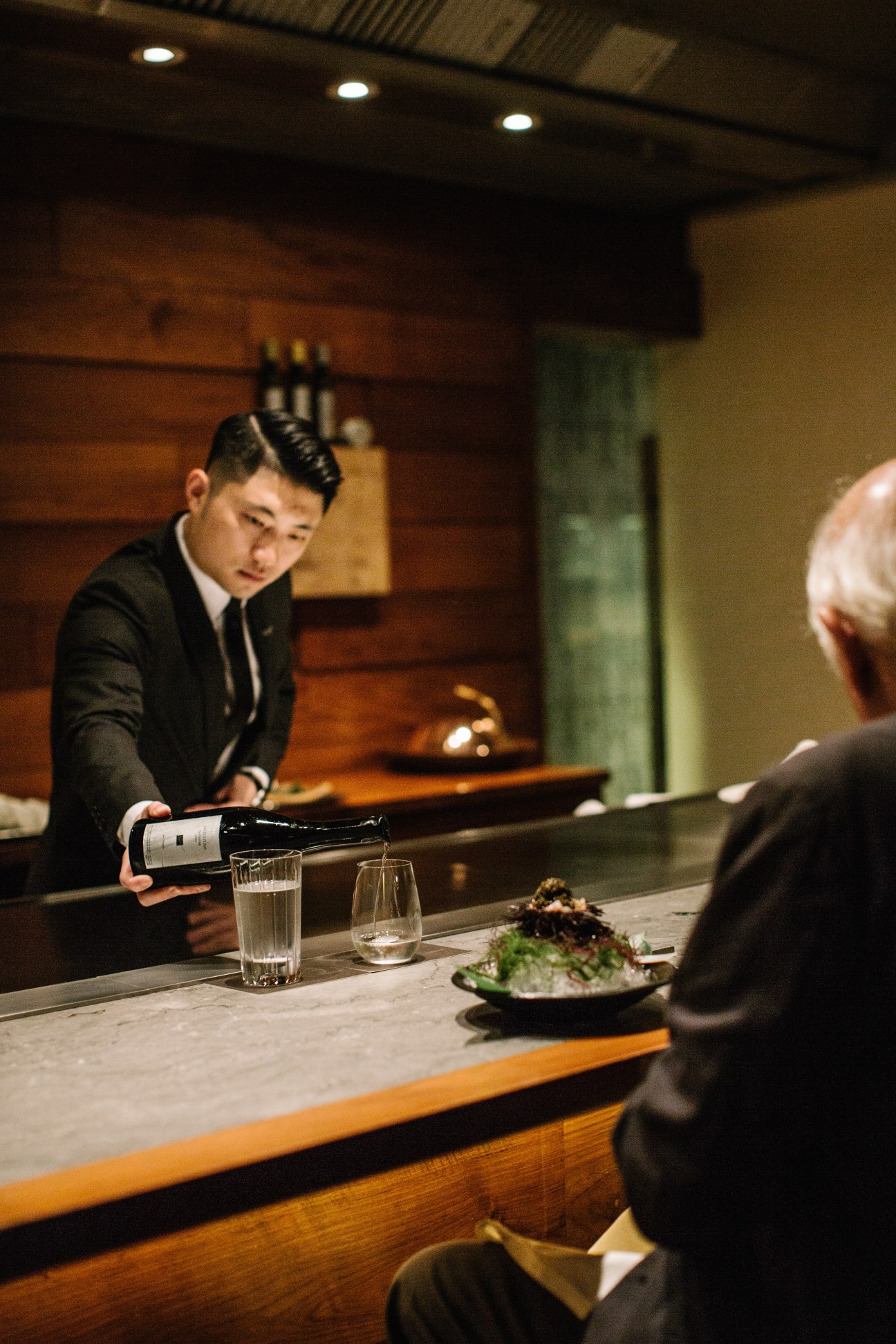 A chef in formal attire pouring Masuizumi Platinum Nama saké at a counter in a Japanese restaurant.