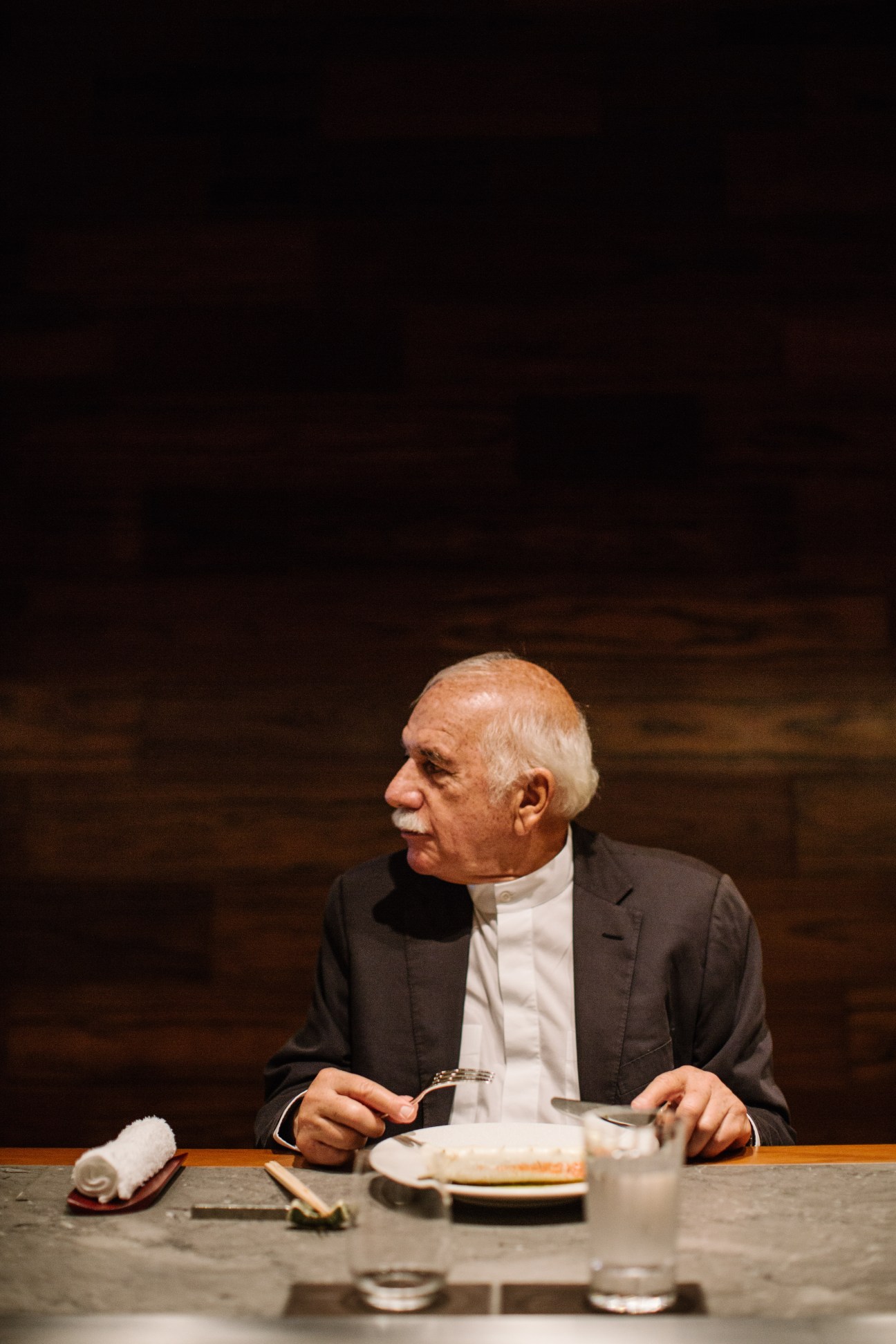 Architect Moshe Safdie seated at a dining counter, holding utensils with a place setting before him.