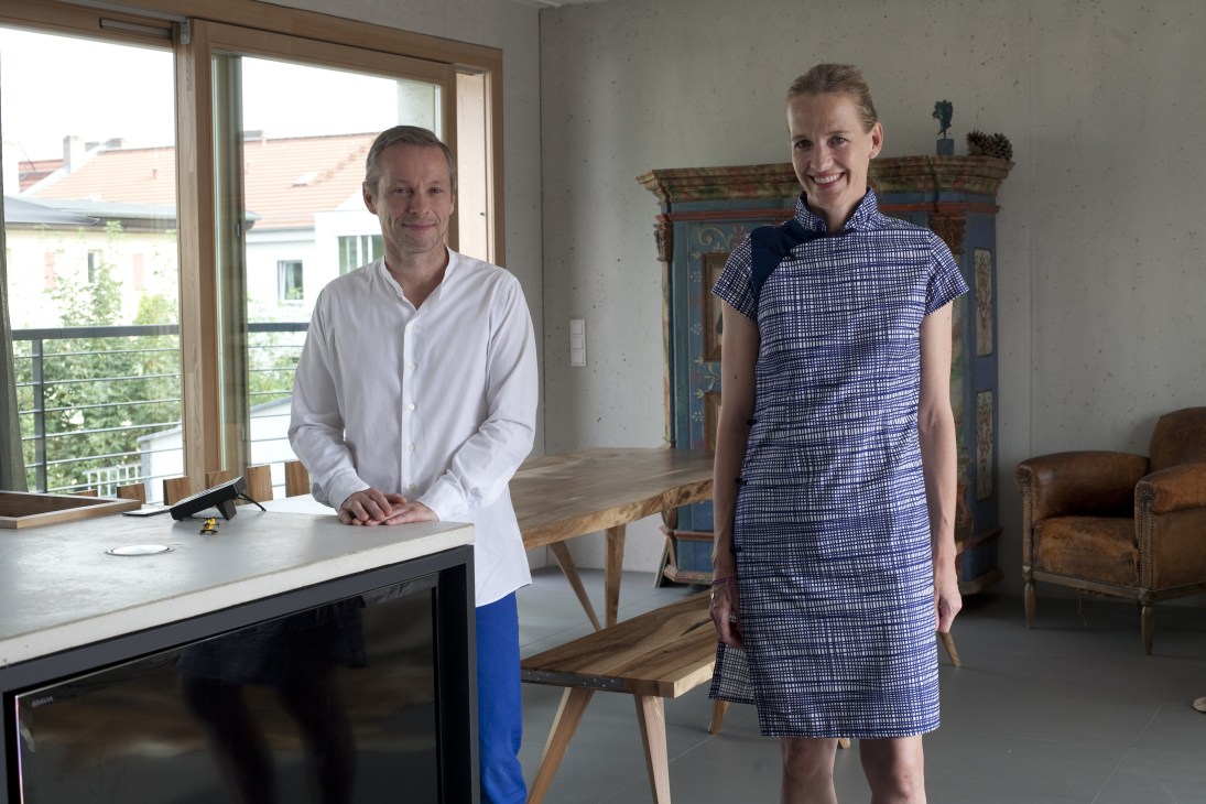 Stefan Karl and Angela Knewitz stand in a bright concrete interior with large windows and wood furnishings.