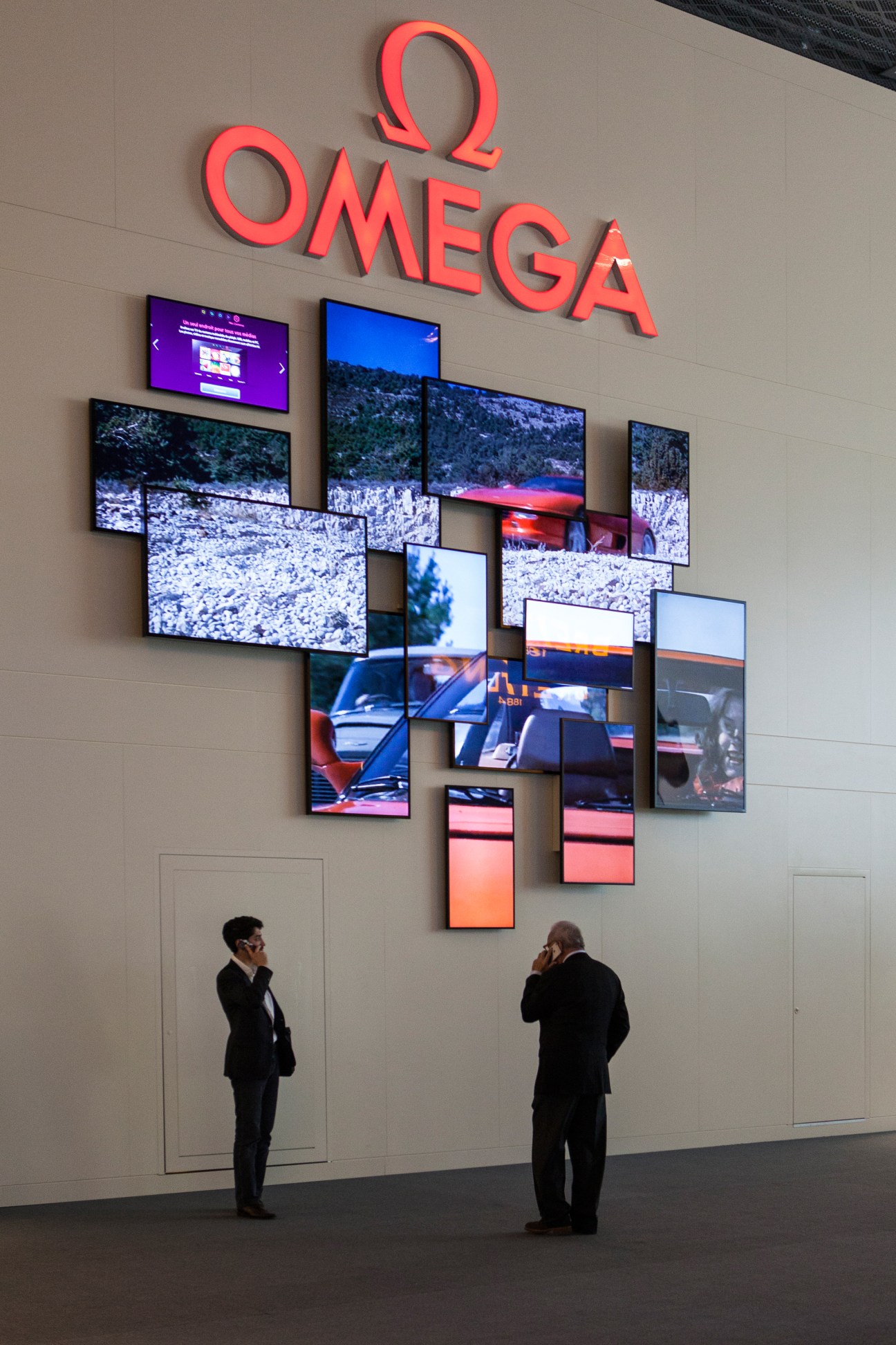 Omega's exhibition stand with illuminated red logo and multiple video screens displaying various content.