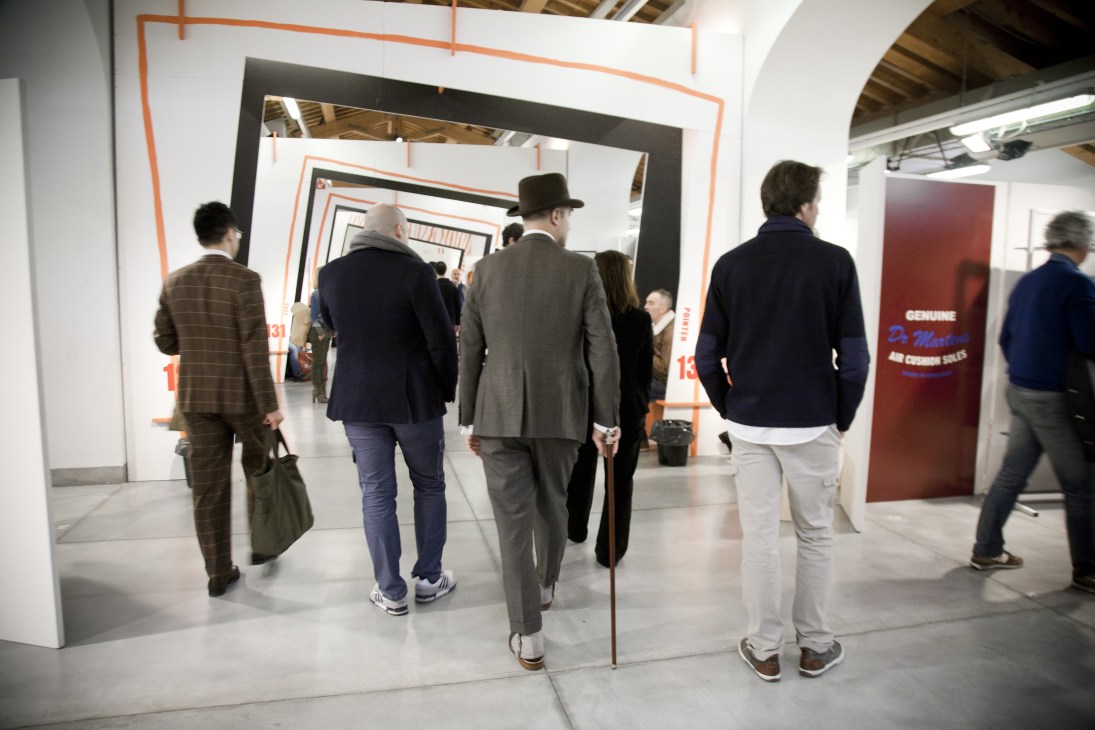 Visitors walking through a modern exhibition booth with orange and black framing at Pitti Uomo menswear tradeshow.