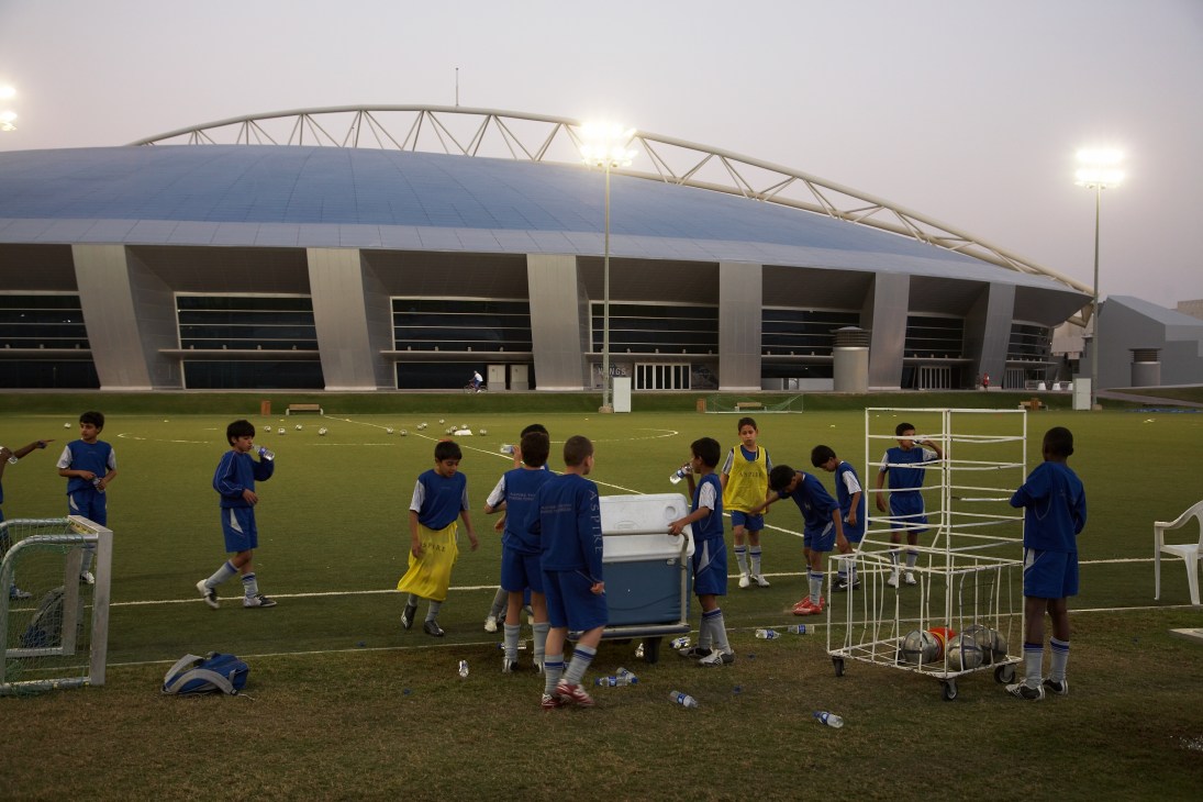 Young football players in blue and yellow training kits practice on a grass field with the Aspire Dome stadium visible in the background.