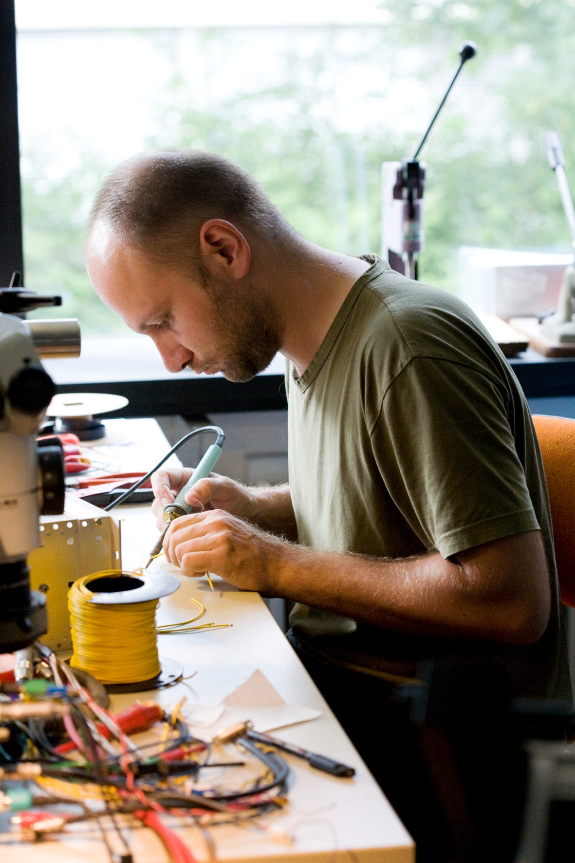 Technician soldering cables at a workbench with electronic components and equipment visible.