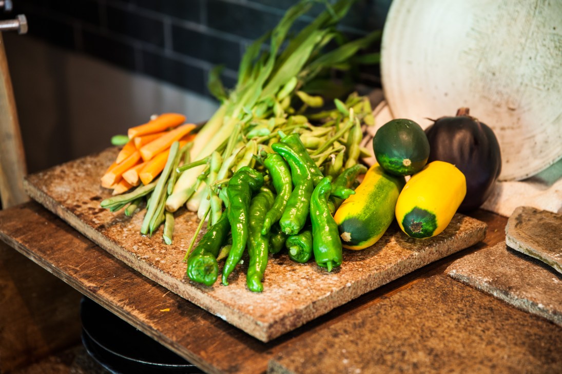 Fresh vegetables including green chilies, yellow peppers, carrots and spring onions arranged on a cork board in a kitchen setting.
