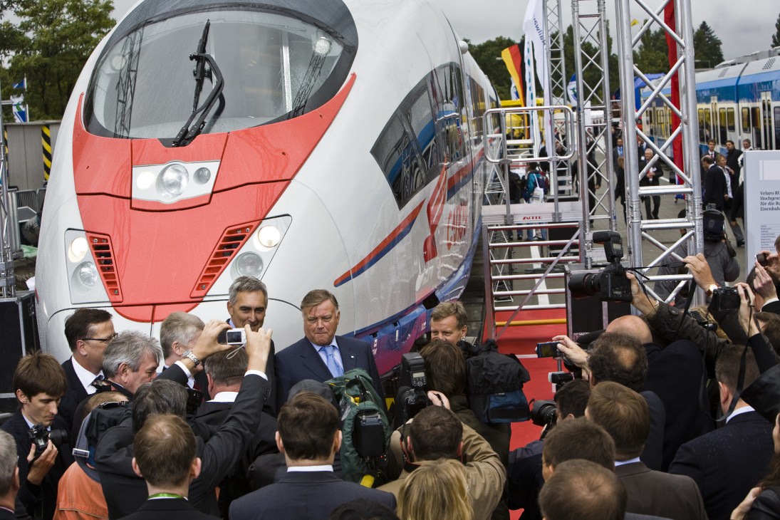 Dignitaries and journalists gather on a red carpet at the unveiling of a red and white Siemens high-speed train at Berlin's InnoTrans trade fair.
