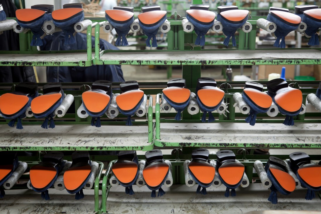 Rows of Santoni shoe lasts with iconic orange soles mounted on green metal shelves in a factory.