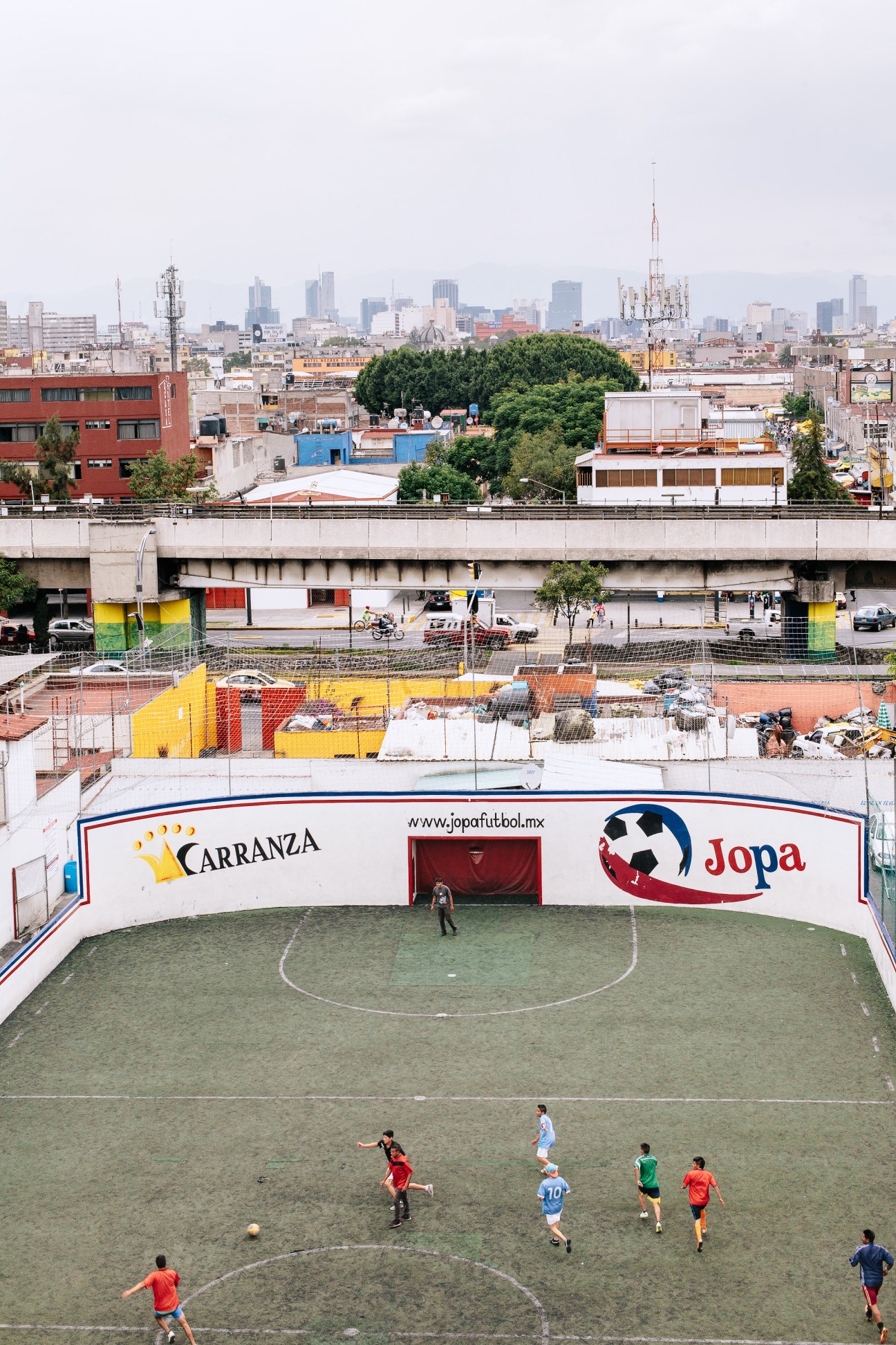 Children playing football on an inner-city pitch with Mexico City skyline in the background.