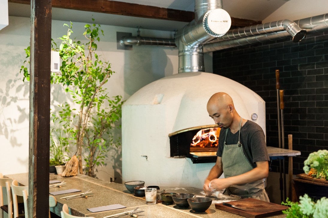 Chef working at counter in Monk restaurant with wood-fired oven visible in background.