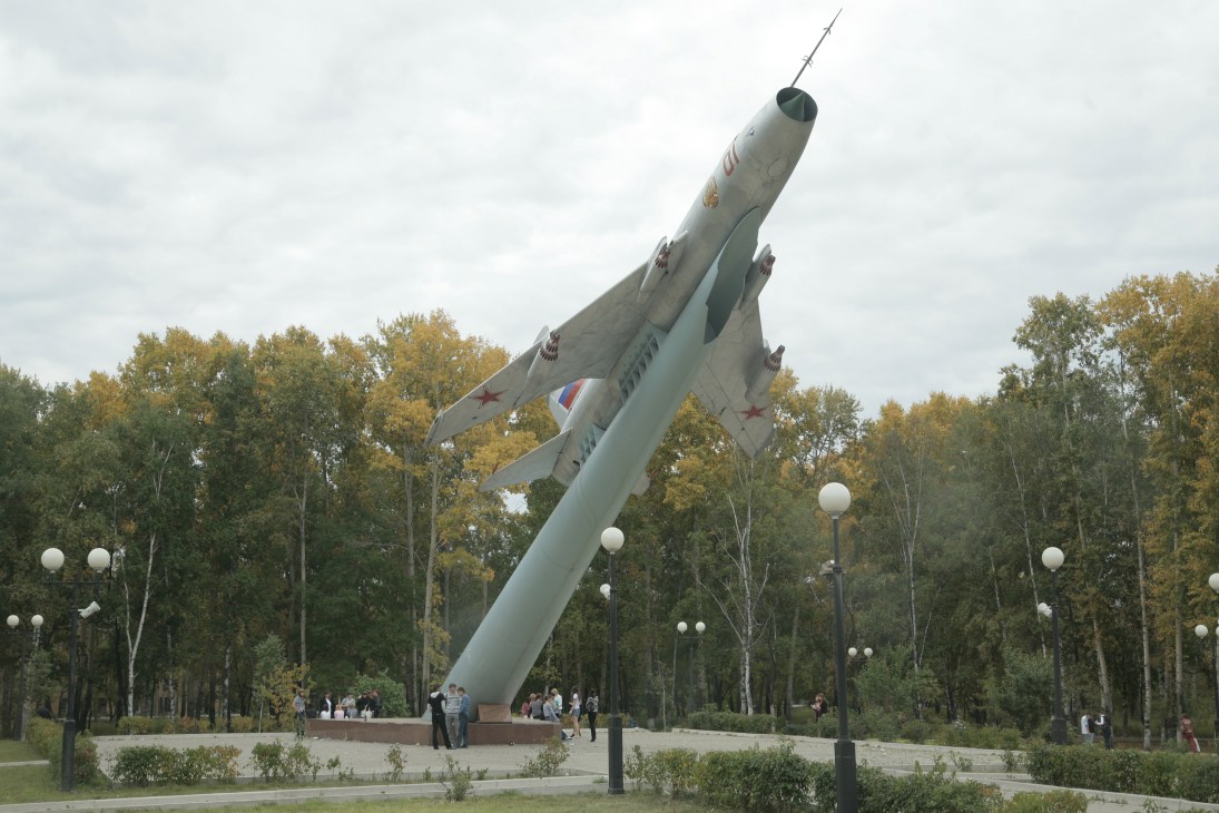 A Soviet-era fighter jet tilted upright on display in a public park with autumn trees and visitors below.