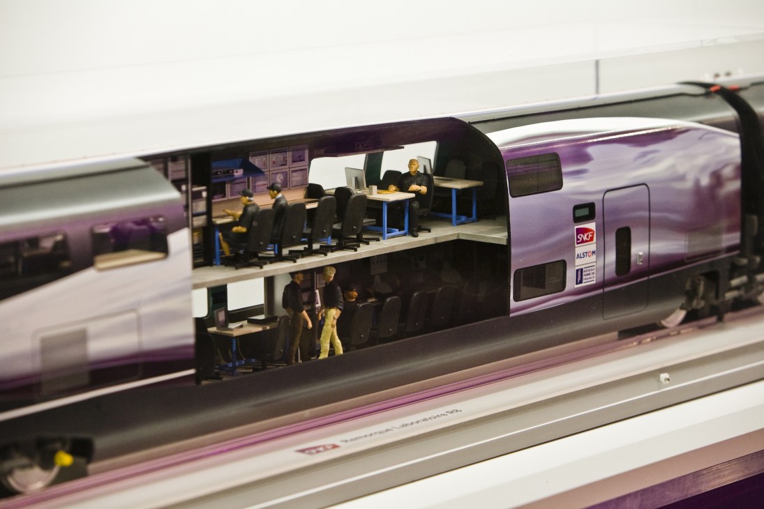 Model train cutaway showing interior layout with passenger seating, work areas, and blue tables.