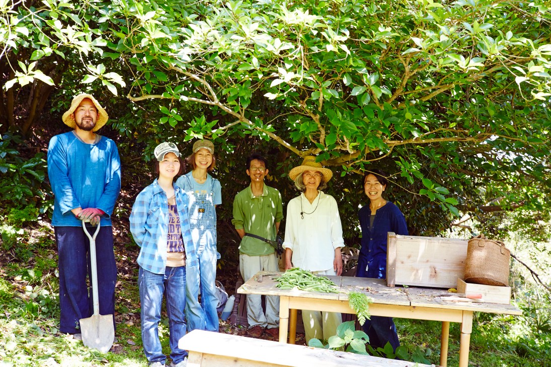 Six people posing together on shared farmland under large green trees in Hayama, Japan.