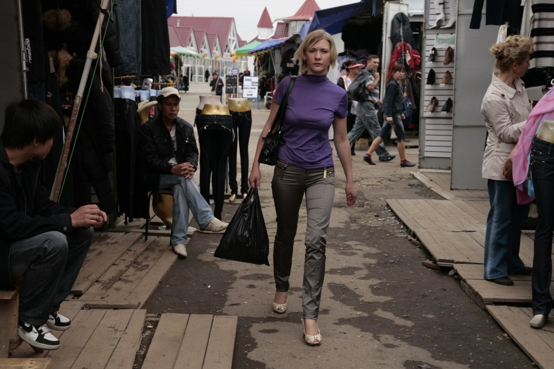 Woman shopping at a suburban outdoor market in Khabarovsk with vendor stalls and other shoppers visible.