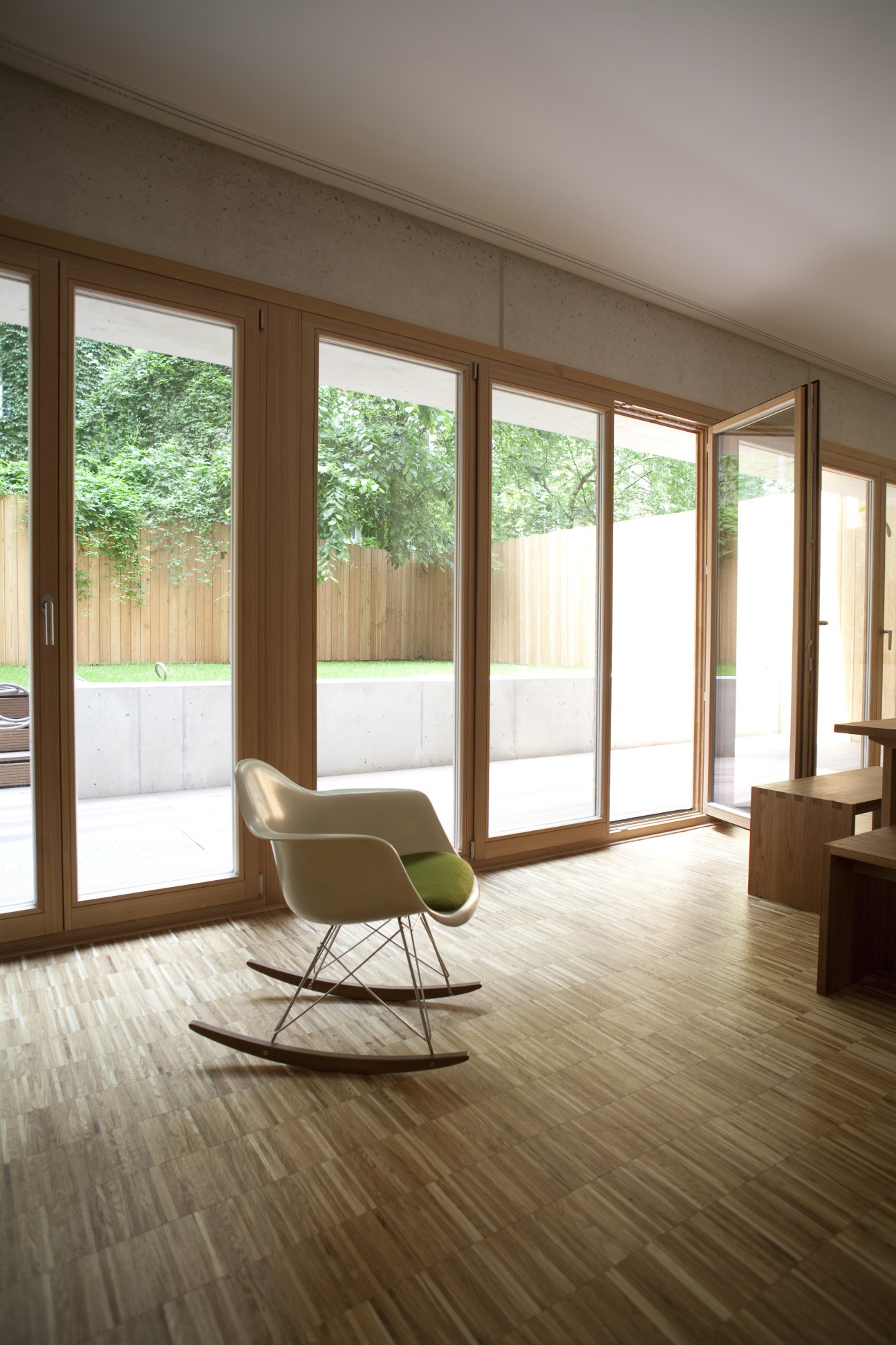 Minimalist concrete interior with wood-framed sliding glass doors opening to a garden, featuring a modern rocking chair.