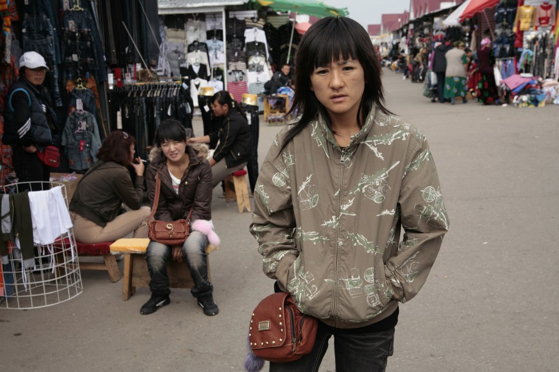 Woman in beige jacket with embroidered designs stands at suburban outdoor market with shoppers and clothing stalls in background.