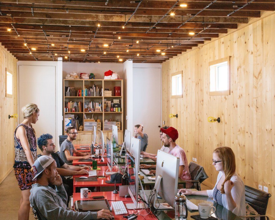Design studio staff working at computers in a light wood-paneled office with exposed ceiling beams and track lighting.