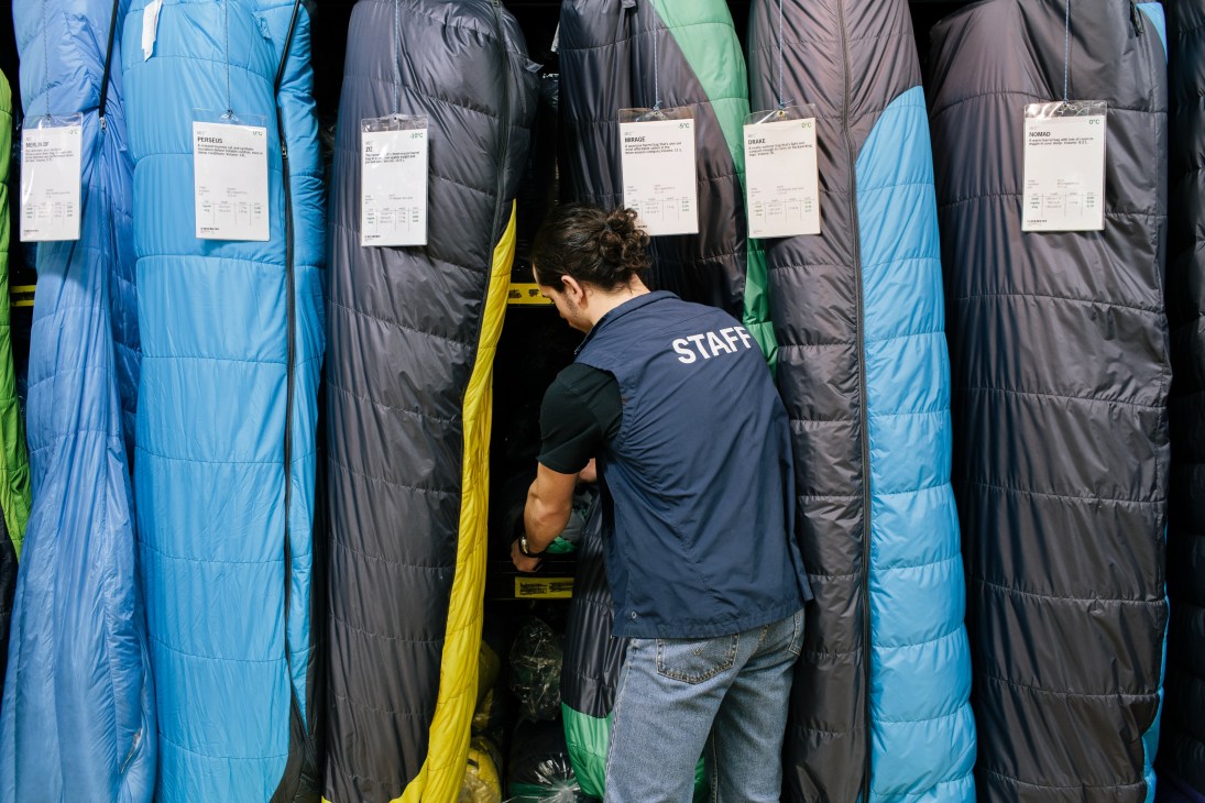 MEC staff member examining sleeping bags displayed in multiple colors with product specification tags.