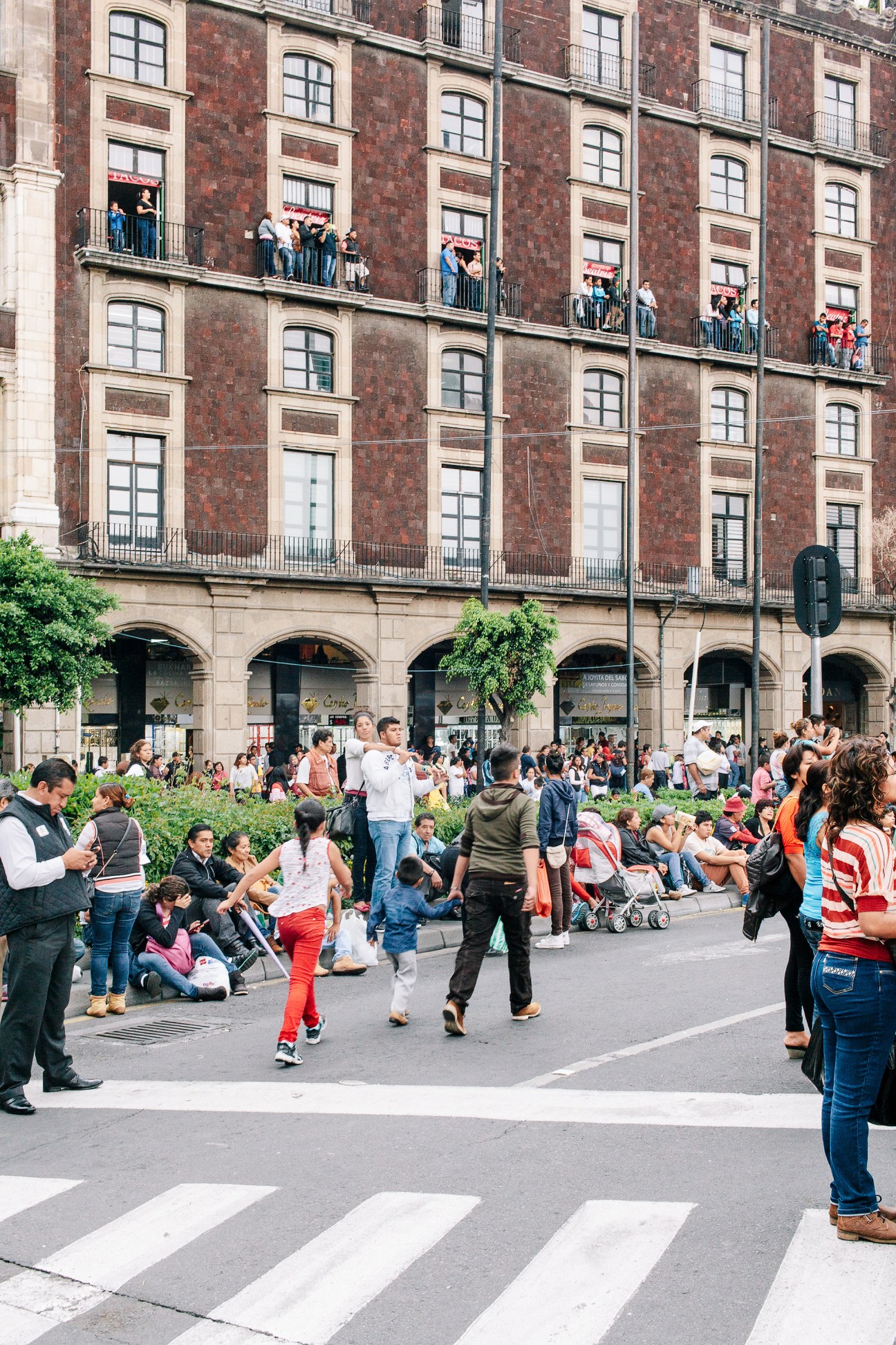 Crowds of people gather in Mexico City's Zócalo main square in front of a large colonial brick building with balconies.