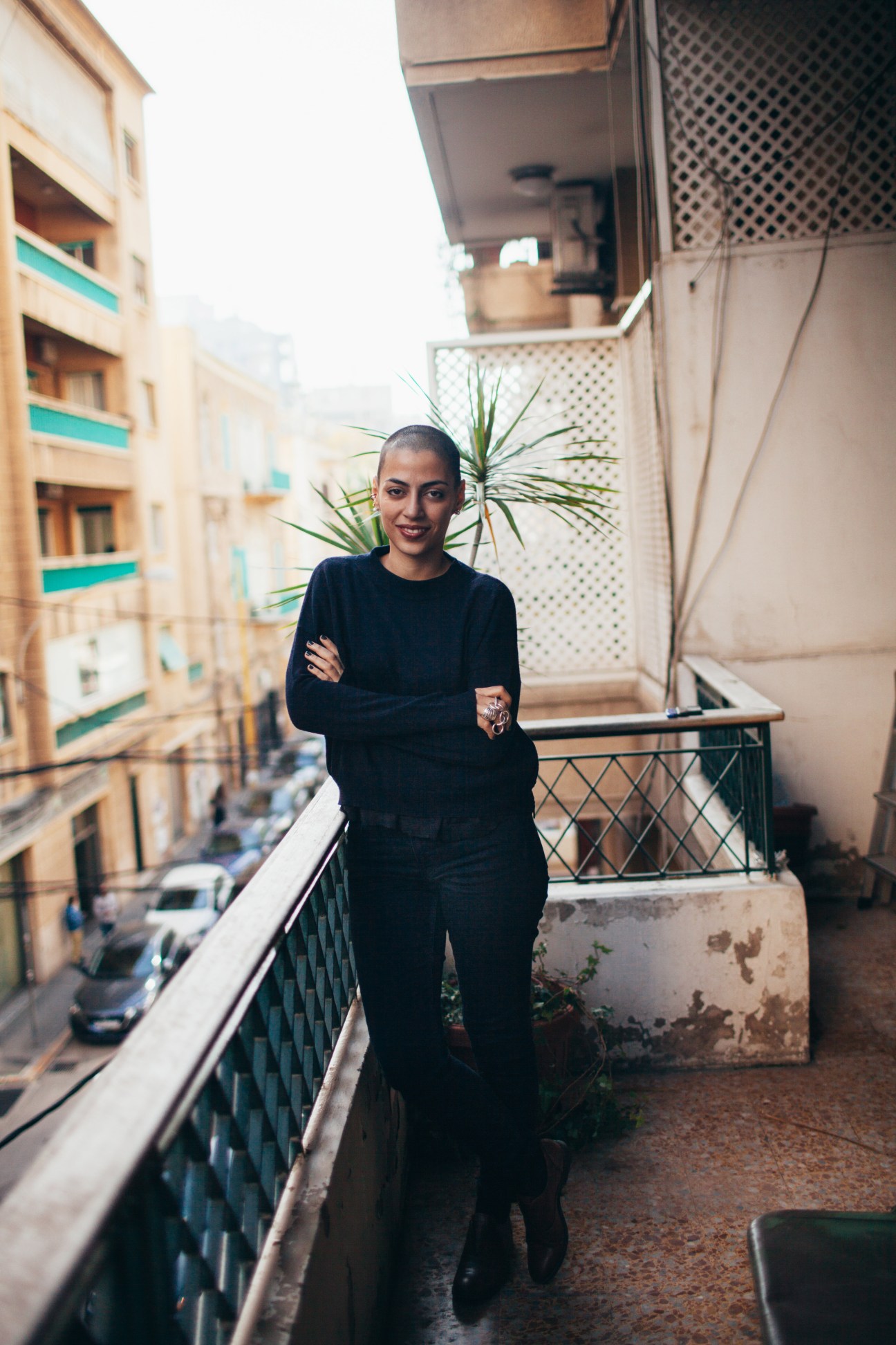 Beirut fashion designer Lara Khoury stands on a balcony overlooking the city's residential buildings, wearing all-black attire and large jewelry.