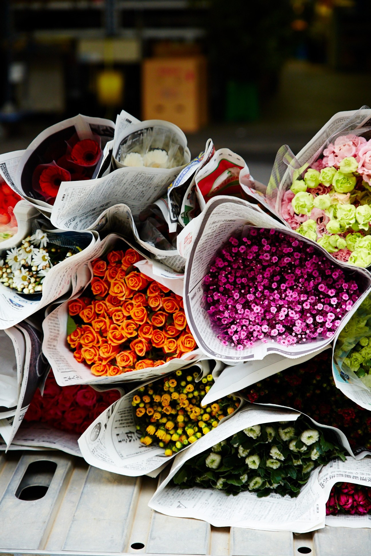 Bunches of colorful flowers wrapped in newspaper, including orange roses, pink chrysanthemums, and yellow flowers at a market.