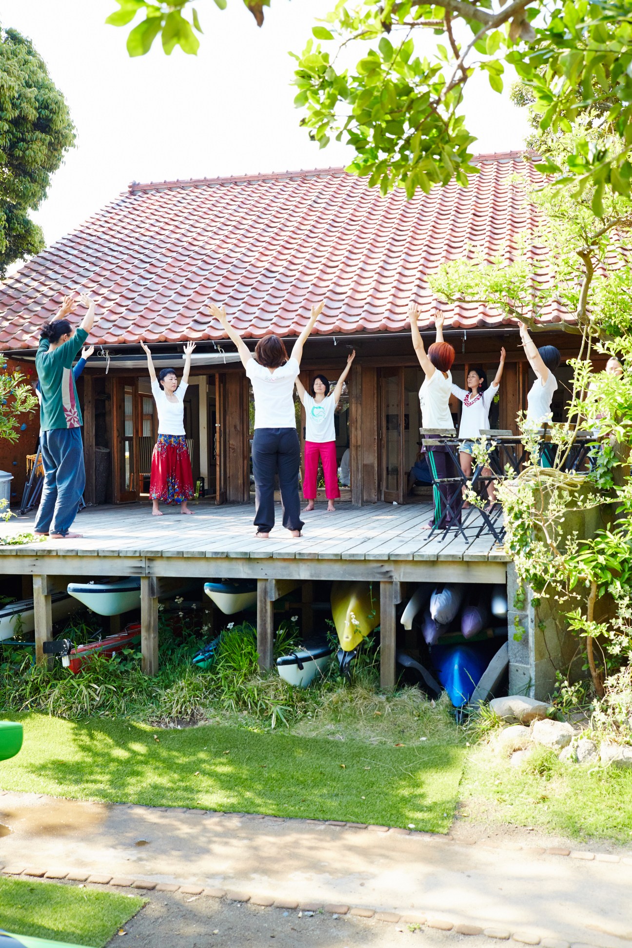 Group of people performing breathing exercises with raised arms on a wooden deck outside a traditional Japanese house.