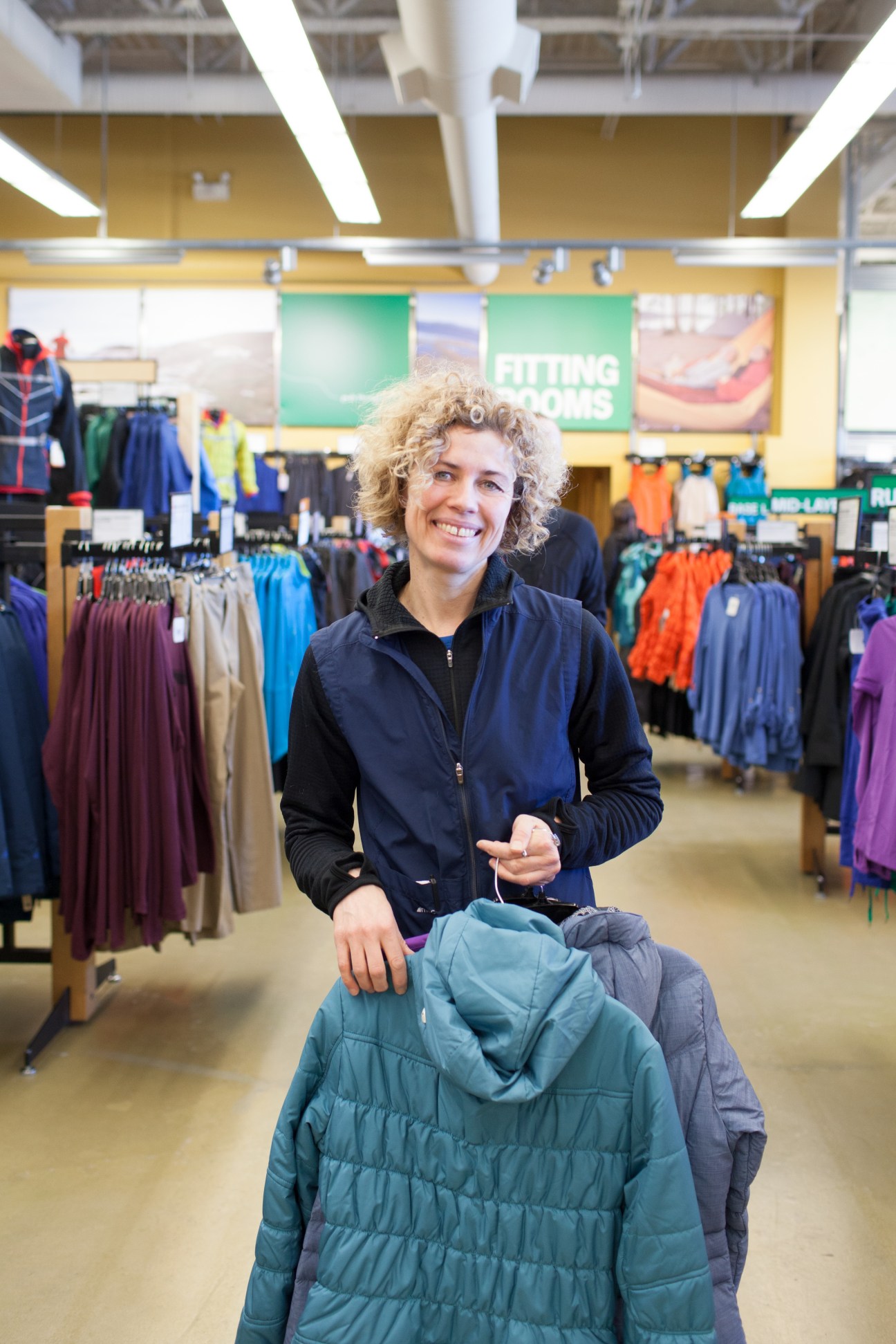 MEC employee Jackie Degoeij holding a teal down jacket in a retail store with outdoor clothing on display.
