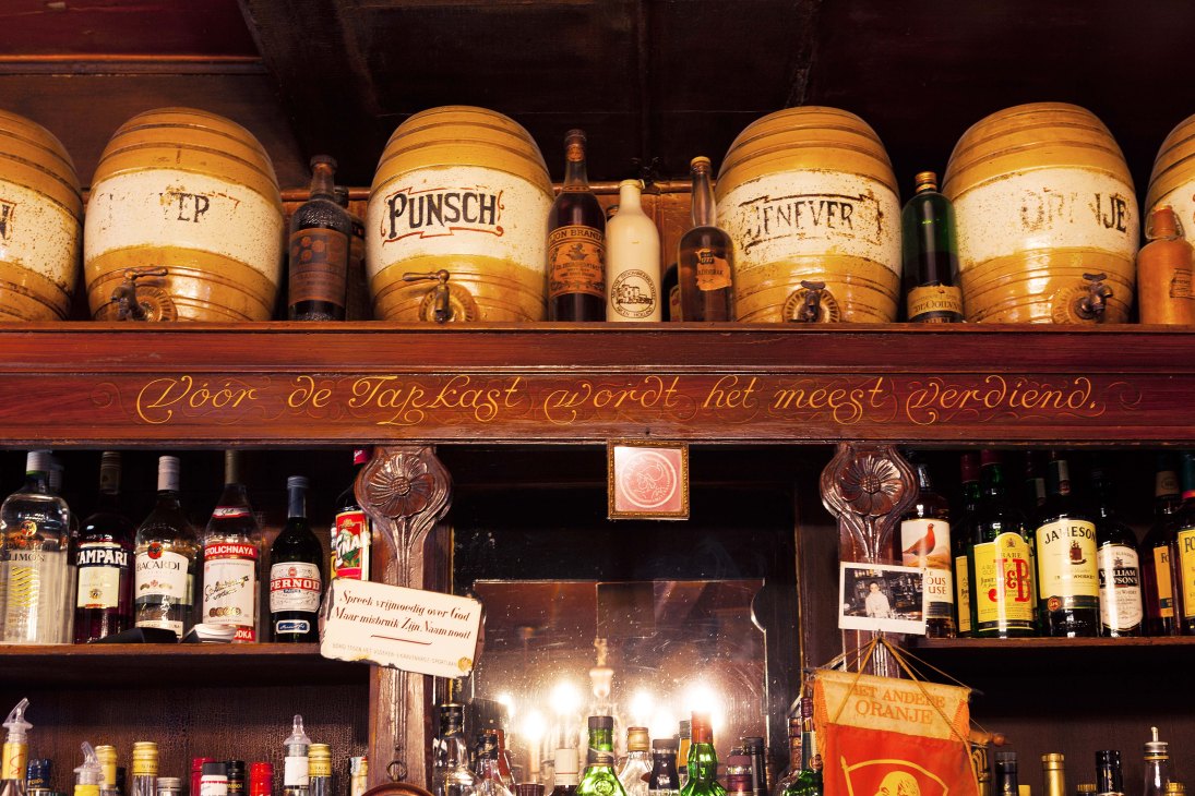 Interior of Café Hegeraad showing wooden shelves with ceramic drink dispensers and bottles, decorated with ornate golden cursive lettering on dark wood.