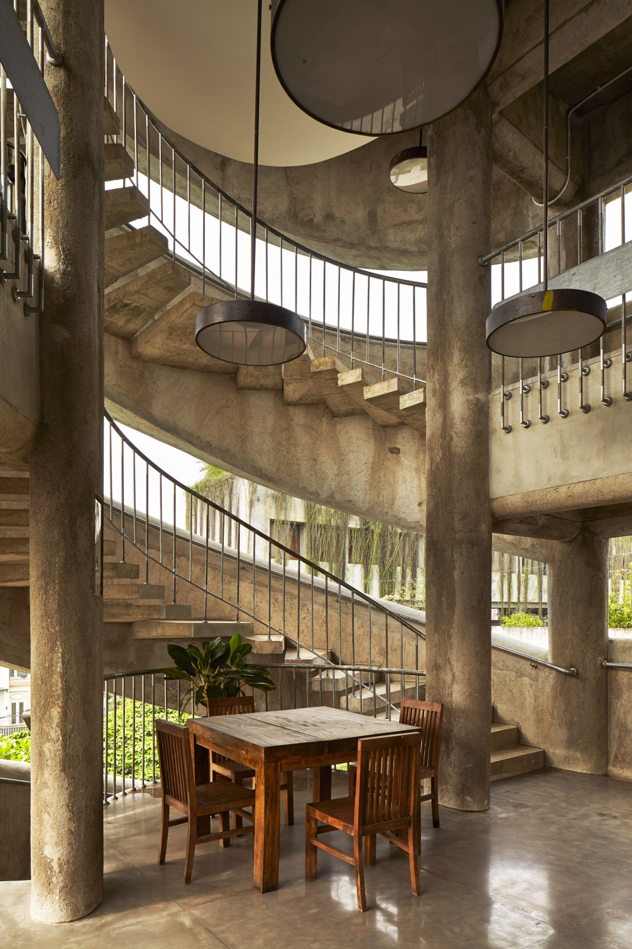 Multi-level concrete interior with curved staircase, ceiling fans, and wooden dining table at Anjung Salihara