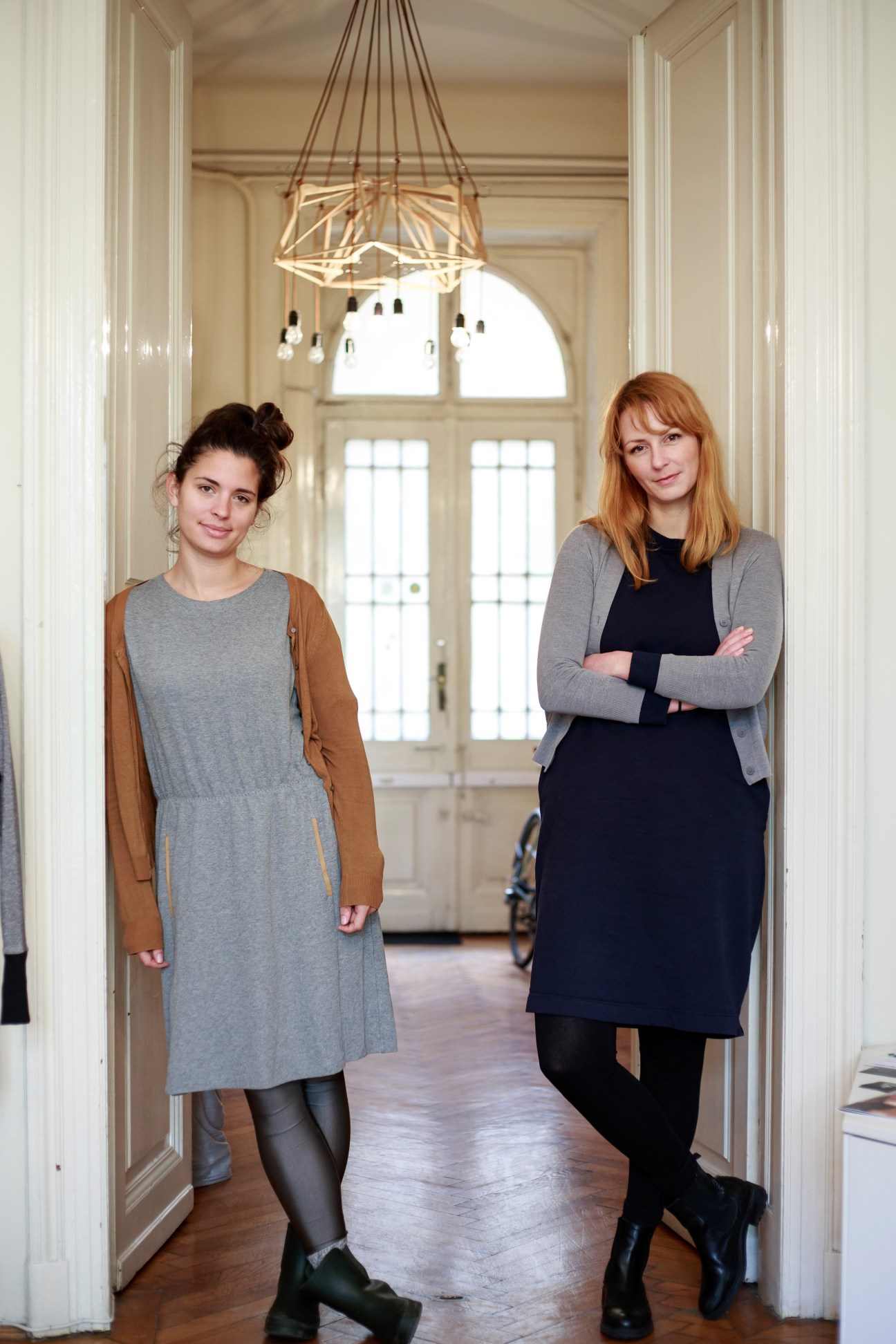 Two women standing in a bright hallway with geometric pendant lighting overhead, one wearing a grey dress with brown cardigan, the other in a navy dress with grey cardigan.