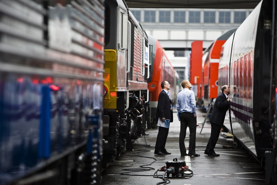 Three men in business attire inspect a red and white Talgo 250 high-speed train at an indoor railway depot.