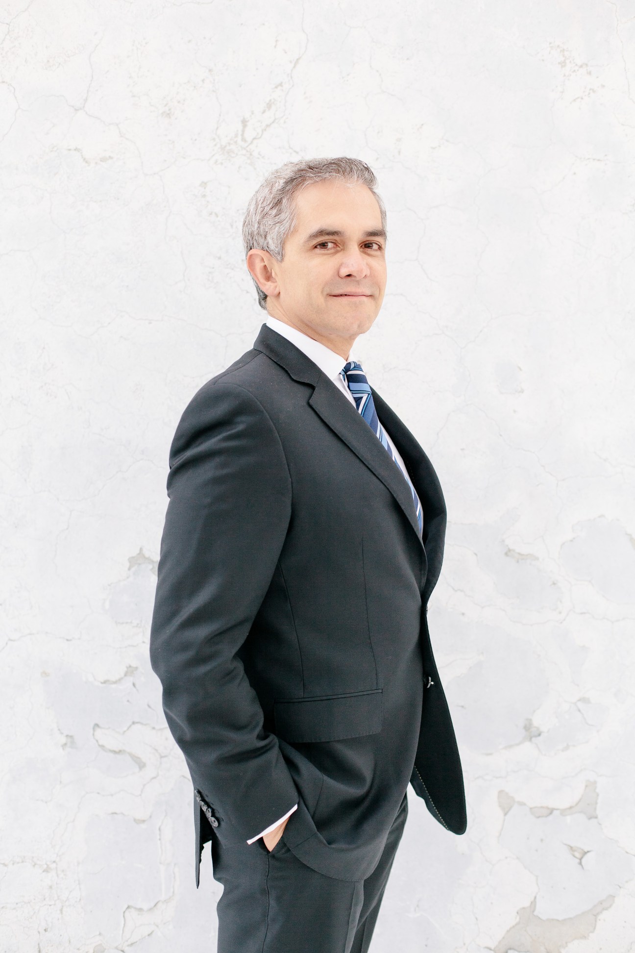 Silver-haired man in dark business suit and blue striped tie standing against white textured background.