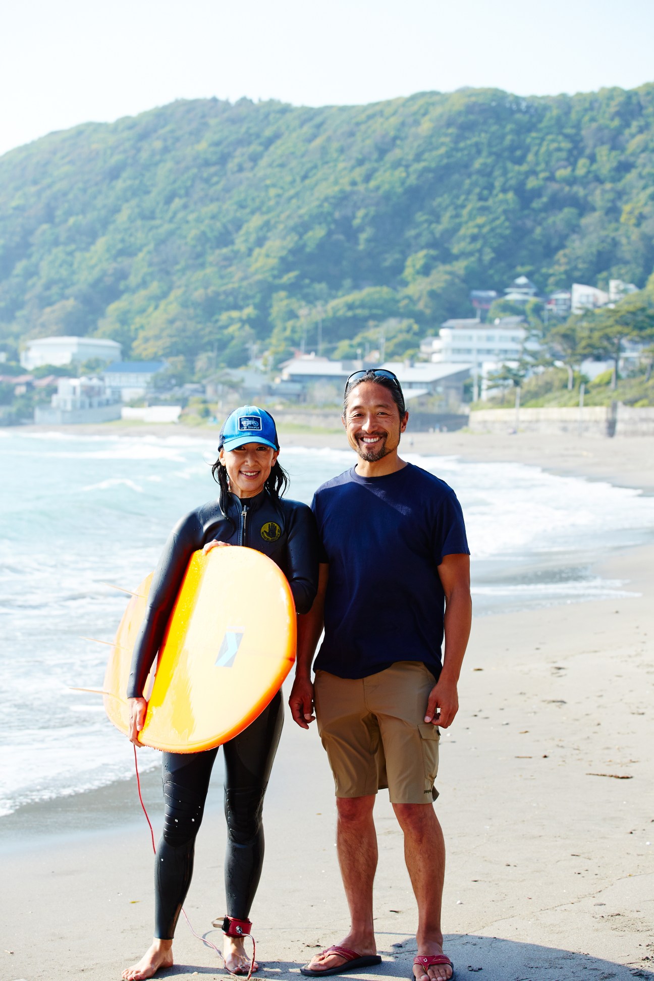 Kazushi Tsuda and wife Minori standing on a beach holding a yellow surfboard, with forested hills and buildings behind them.