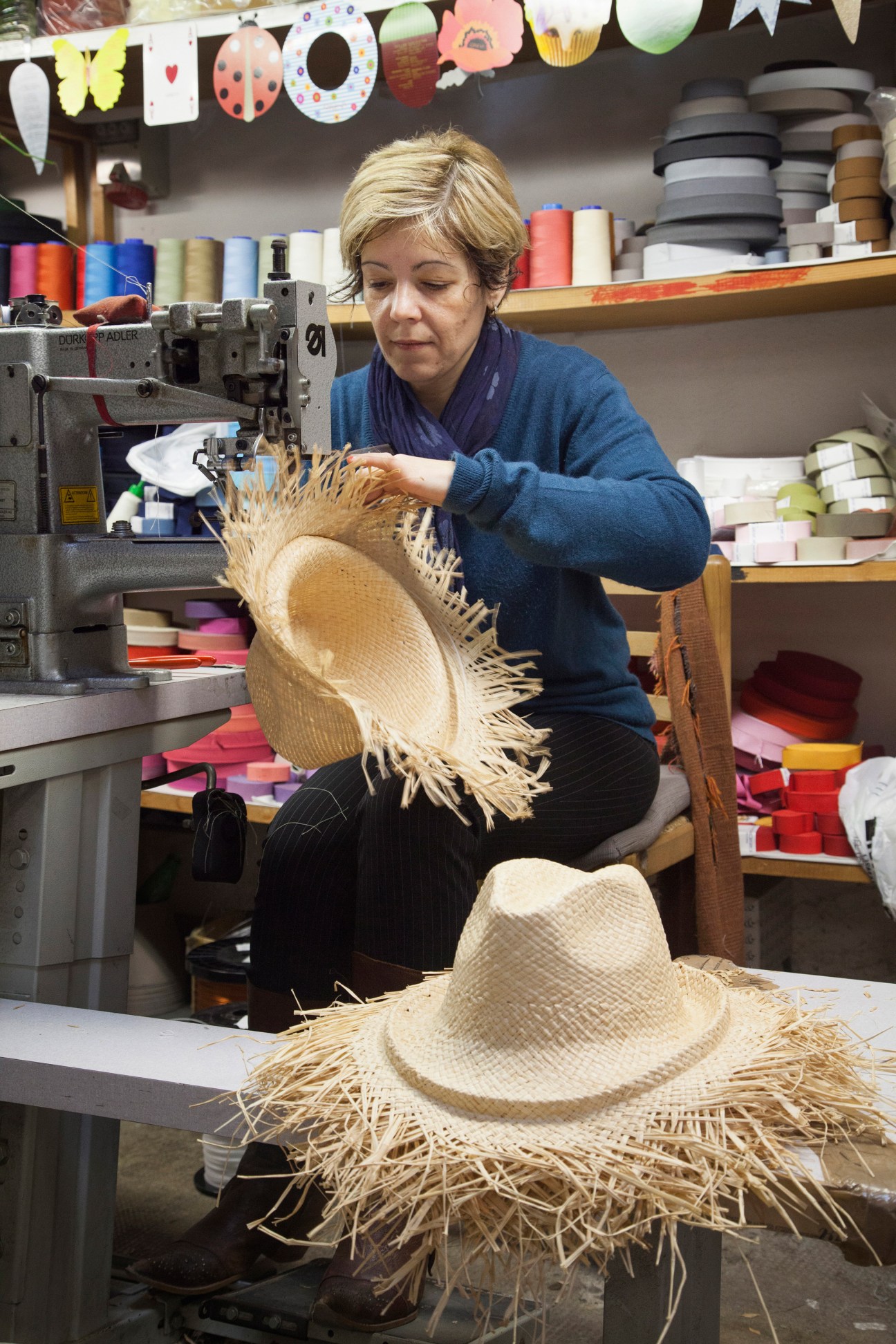 Seamstress operating a vintage sewing machine to stitch the brim of a woven straw hat in a colorful workshop.