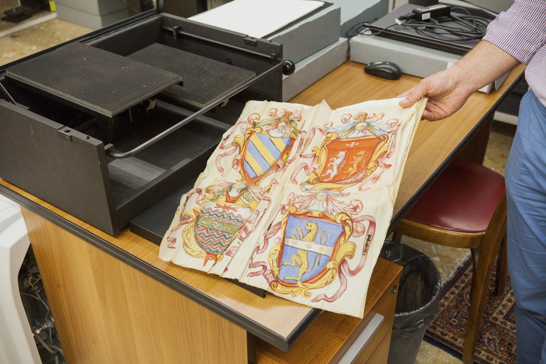 A person's hand holding historic illuminated coats of arms with ornate decorative borders and heraldic shields in gold and color.