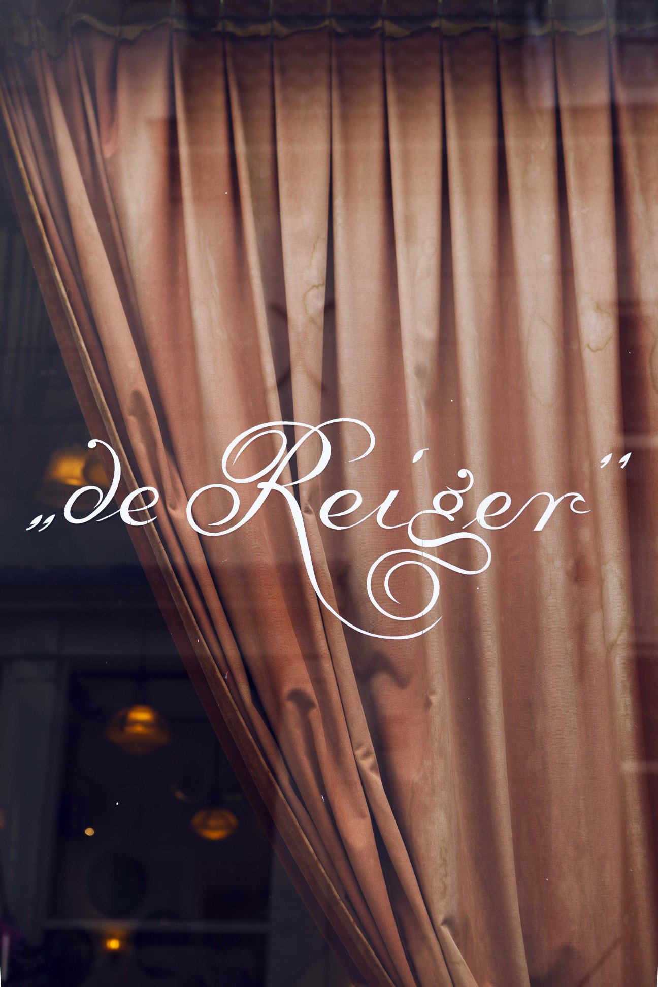 White cursive lettering reading "de Reiger" painted on a window with draped curtains behind it.